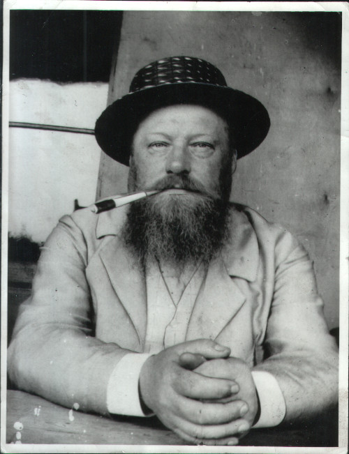 Black and white photograph of Hans Richter, seated with his hands clasped and resting on a table, looking directly into the camera. He is wearing a pale shirt, waistcoat and jacket, with a hat. He also has a cigarette in holder held to to side his mouth. 