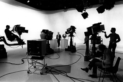 Alt text: Black and white photograph of a television studio. A presenter is being filmed, with camera crew and equipment in the foreground. Cables and lighting are visible.