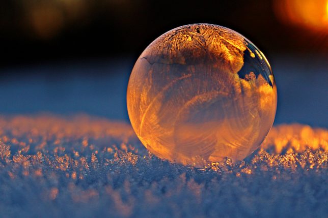 Colour close-up photograph of a clear glass ball, with ice patterns, resting on heavily frosted ground, illuminated by sunlight.
